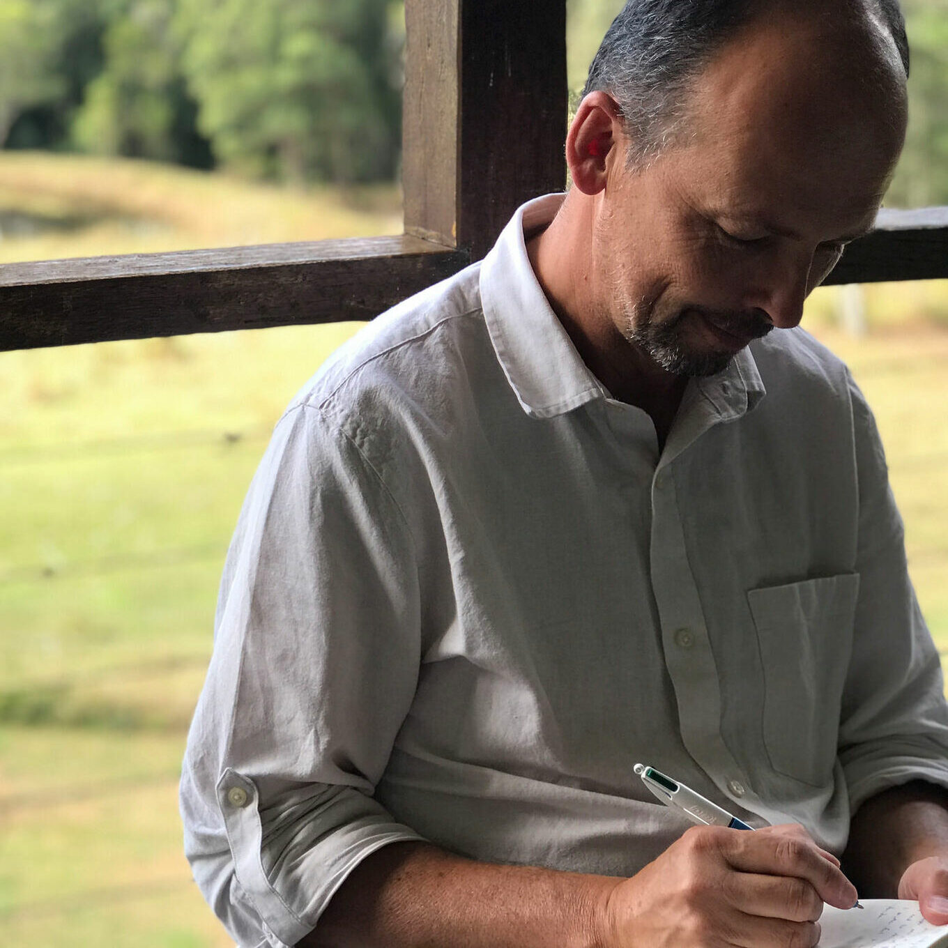 tenneson woolf in light grey shirt looking down and writing with pen on paper in front of brown fence and green pasture