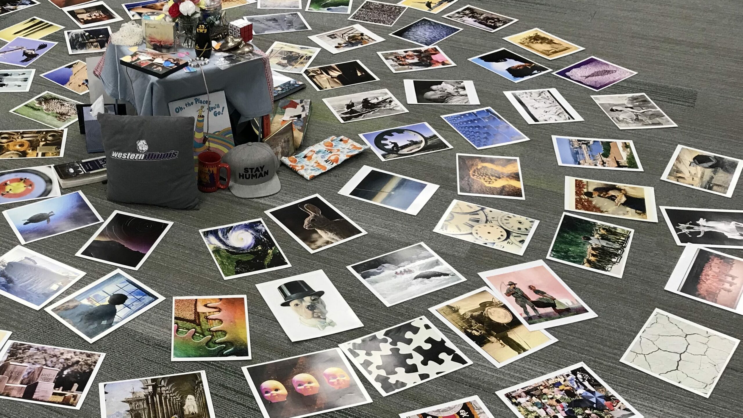group facilitation activity radial display of varied photographs laid flat on on grey carpet floor including western illinois gear a hat that says stay human and a copy of the dr suess book oh the places you'll go